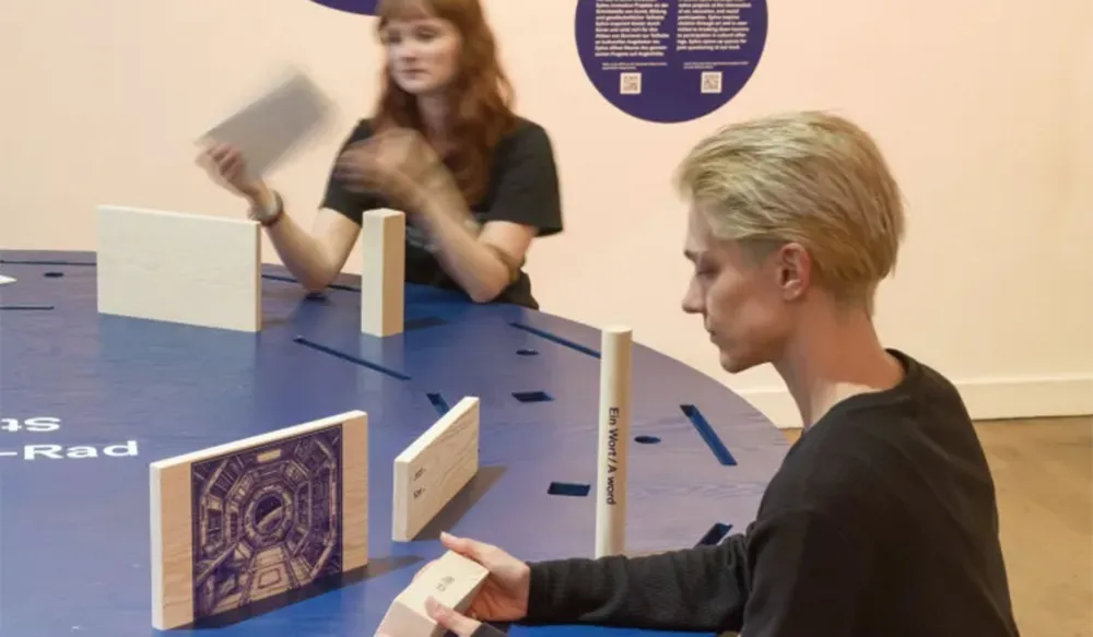 Two people are sitting at a round table in an exhibition space, looking at wooden objects.
