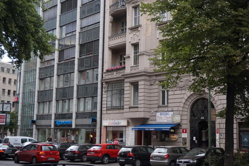 Color photo: Façades of a prewar and a postwar Berlin building, with trees and cars
