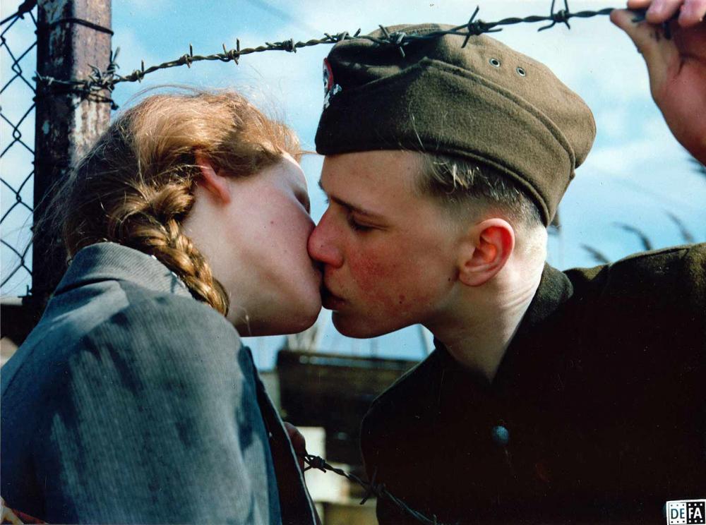 Film still: a girl with blond pigtails and a boy in Nazi uniform kiss through a barbed wire fence.