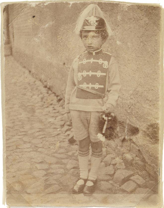 Photography of Walter Frankenstein as a child, costumed on cobblestones standing in front of a house wall