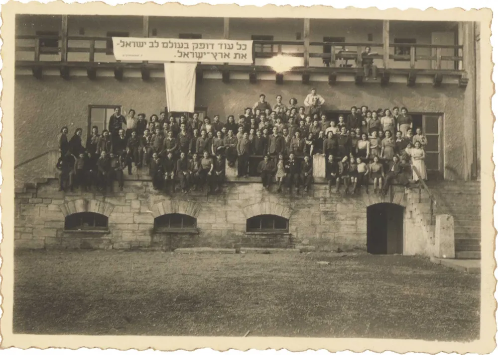Black and white photography. About 100 women and men sit on a porch. Above them hangs a banner with a Hebrew quote from a song by Pooa Grinshpon.