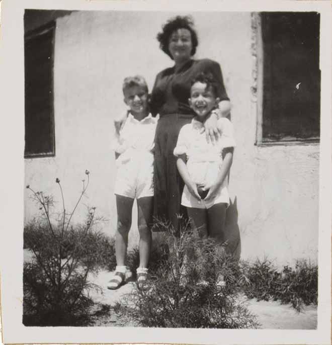 Auf der Schwarz-Weiß-Fotografie legt Leonie im dunklen Kleid ihre Hände auf die Schultern der links und rechts stehenden und in weißen Shorts und Hemden gekleideten Kinder. Im Hintergrund ist eine Hauswand zu sehen.