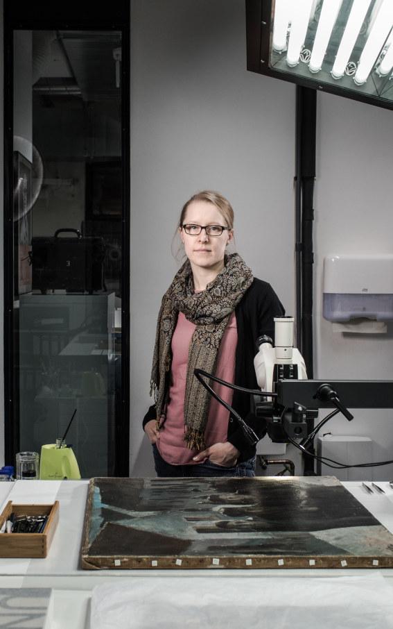 Eine junge Frau steht an einem Arbeitstisch in einem Atelier, vor ihr liegt ein altes Ölgemälde.