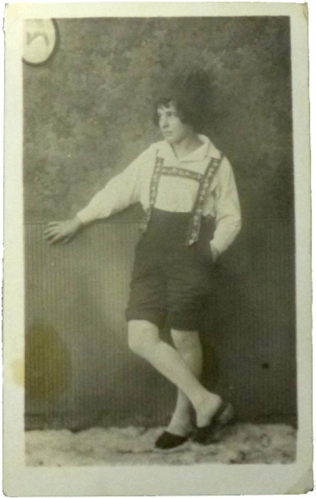 Studio portrait of an unknown woman in full-length figure with slippers, light pantyhose, light shirt, dark trousers with decorated suspenders and dark hat