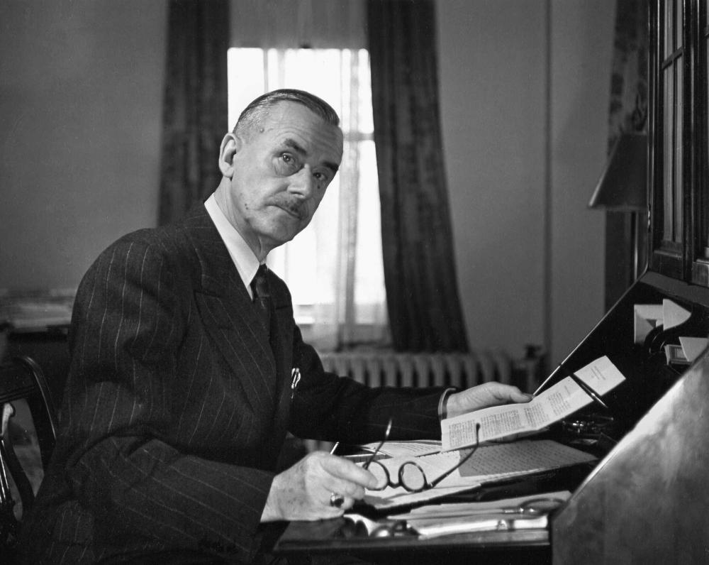 Black-and-white portrait of Thomas Mann, sitting at a writing desk with his glasses and paper in hand, looking at the camera
