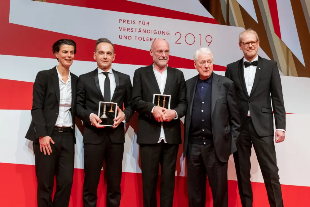 Two men with prizes in their hands between the laudators and the director.