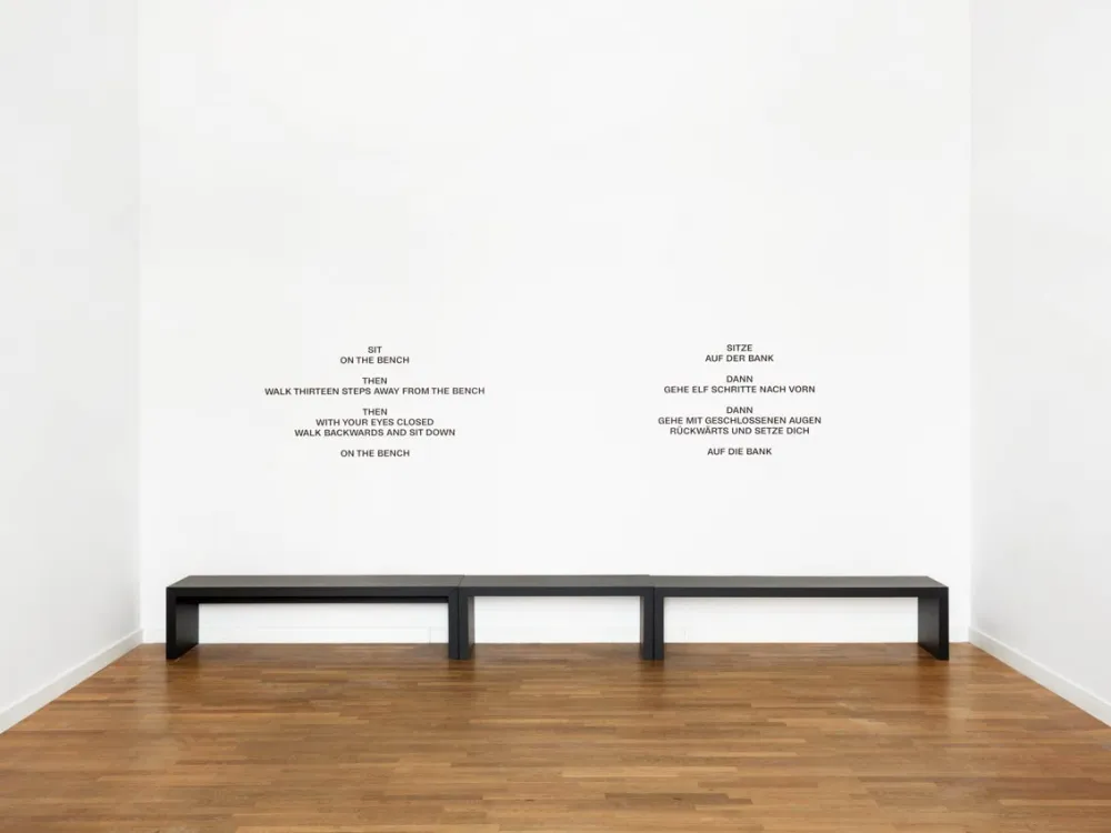 Eine breite schwarze Sitzbank in einem wei&szlig;en Ausstellungsraum, an der wei&szlig;en Wand dahinter Handlungsanweisungen, wie man sich von der Bank weg und wieder zur&uuml;ck bewegen soll.