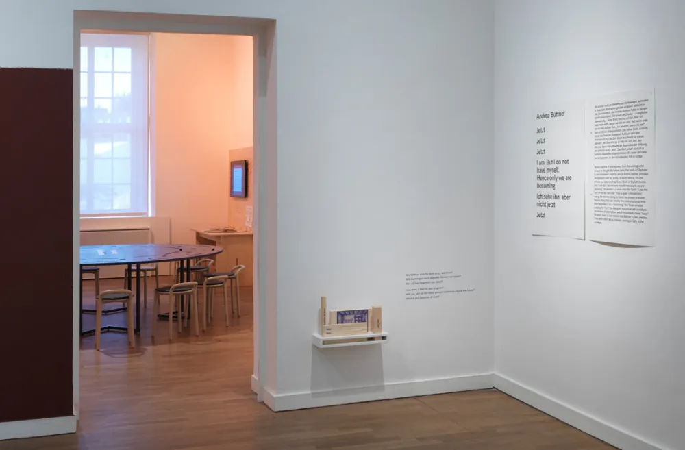 Blick in die Ecke eines Ausstellungsraums, wo auf niedriger H&ouml;he Holzelemente zum Herausnehmen angebracht sind und daneben Fragen an der Wand stehen. Durch eine T&uuml;r&ouml;ffnung sieht man im Nebenraum einen runden Tisch mit St&uuml;hlen und eine interaktive Medienstation.