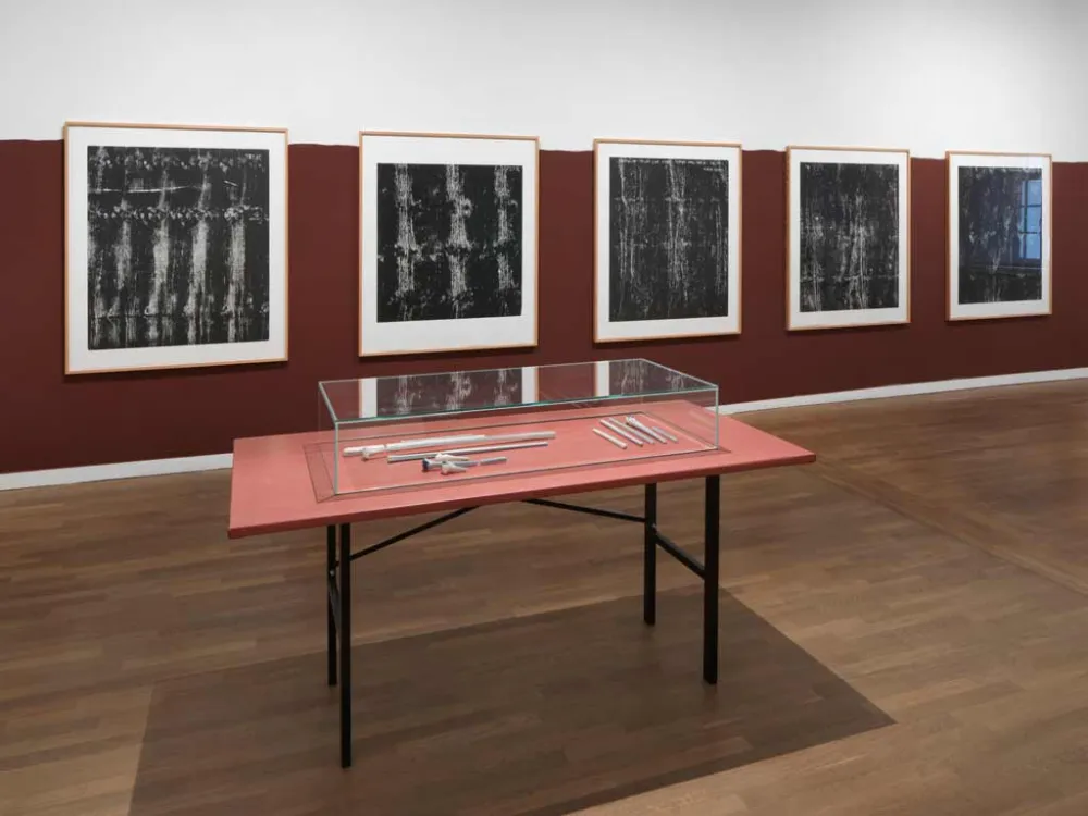 A view into an exhibition room featuring five black-and-white woodcuts, framed against a wine-red wall; in front of them is a table with a glass display case containing bone-shaped glass sculptures.