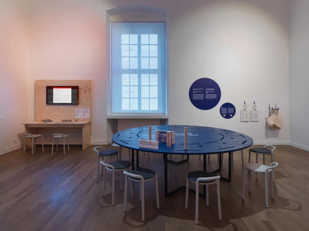 A view of an exhibition space featuring a large round table surrounded by stools, with recesses in the table for inserting wooden pieces. In the background is another interactive station with a screen and tablets.