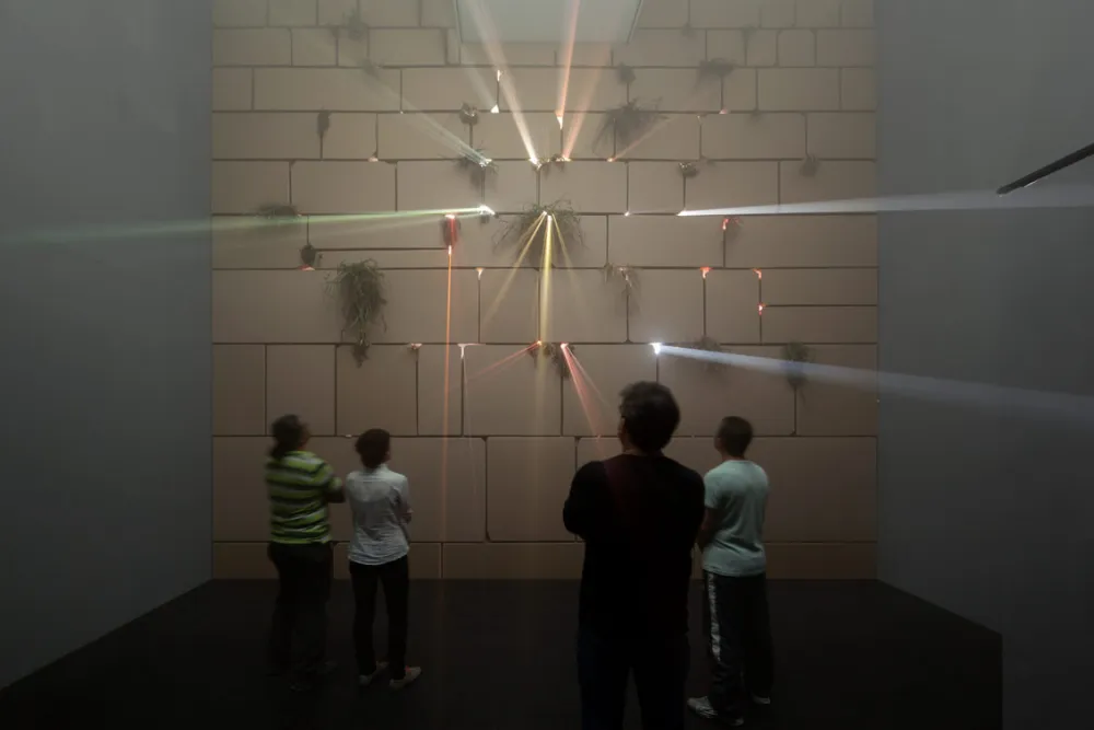 Aus Pappkartons nachgestellte Klagemauer in einem Ausstellungsraum, durch deren Ritzen Diskolicht dringt, davor Menschen, die die Wand anschauen.