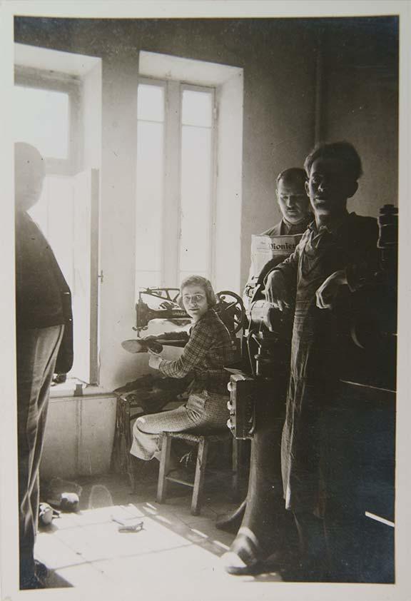 Black-and-white photo of a workshop. A woman sits at a sewing machine in the rear with one man standing to her left and two to her right.