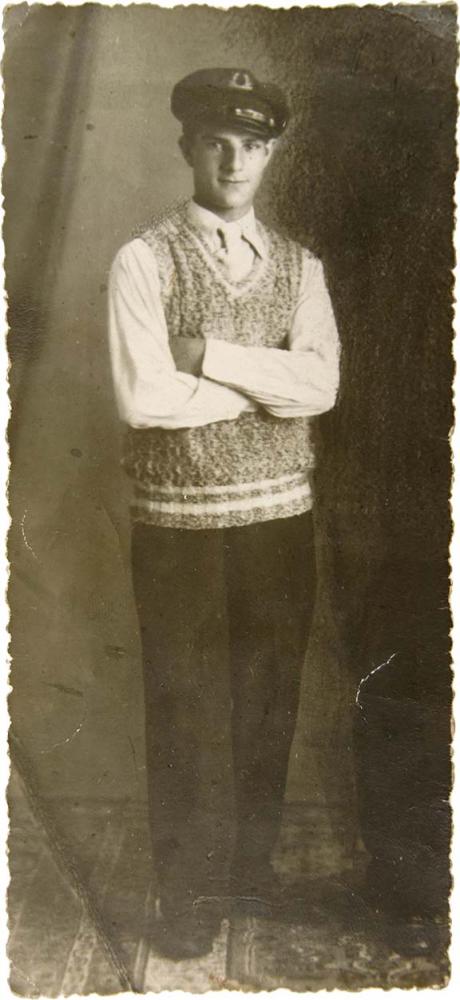 Porträt eines stehenden jungen Mannes, der eine Matrosenmütze und einen handgestrickten Pullunder trägt. Er schaut freundlich in die Kamera und wirkt sehr zerbrechlich