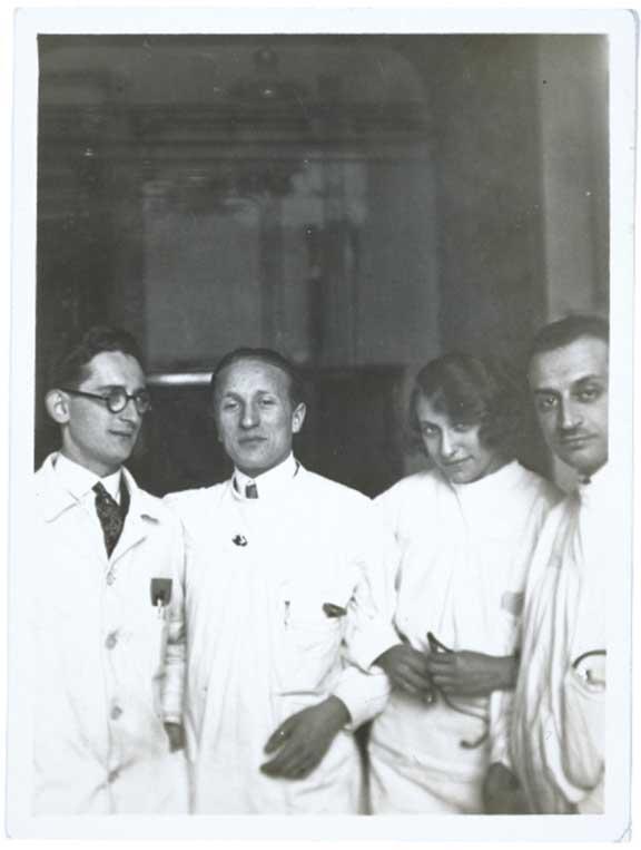 Black-and-white photograph. Erich Simenauer is the second from the left in a group of four. Standing to his right is a woman with a coy expression. Everyone is dressed in white doctor’s coats. None of the three others have been identified.