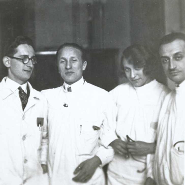 Black-and-white photograph. Erich Simenauer is the second from the left in a group of four. Standing to his right is a woman with a coy expression. Everyone is dressed in white doctor’s coats. None of the three others have been identified.