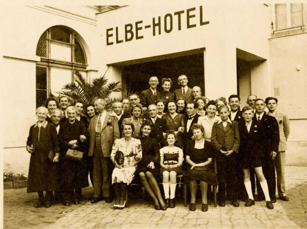 Schwarz-weiß-Fotografie von einer Gruppe, die vor einem Gebäude mit der Aufschrift Elbe-Hotel steht und sitzt.