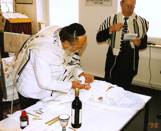Älterer Mann mit Tallit beschneidet ein Baby, das auf einem Tisch liegt. Im Hintergrund steht ein weiterer Mann mit Tallit