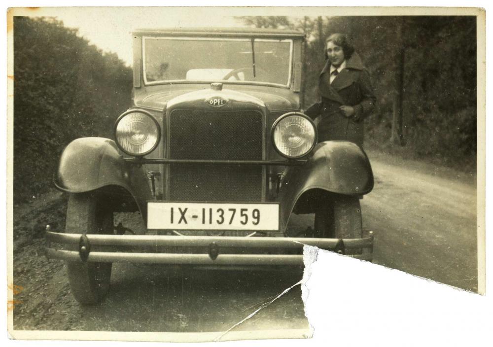 Black and white photo of a young woman in winter clothes next to a car on a country road, the lower right corner of the photograph is torn out.