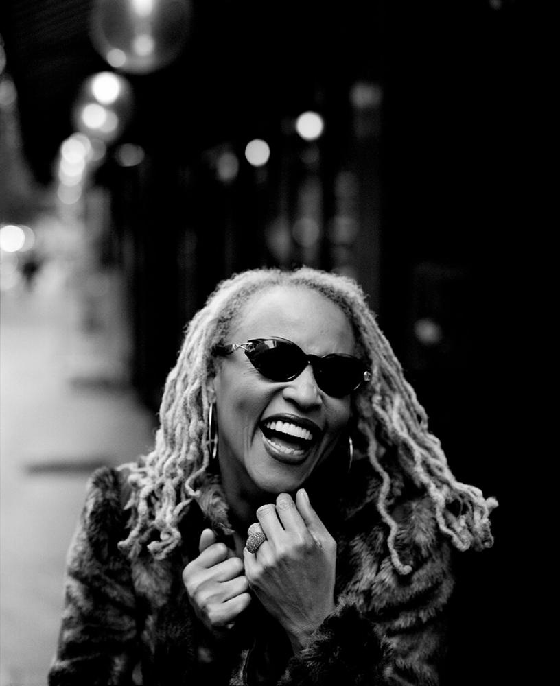 Cassandra Wilson mit Sonnenbrille und Pelzmantel lachend auf einer Straße. 