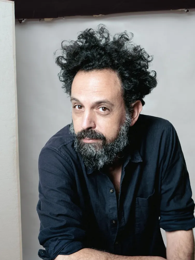 A man with black curls, a mottled black and gray beard and a slightly unbuttoned blue shirt looks animatedly into the camera.