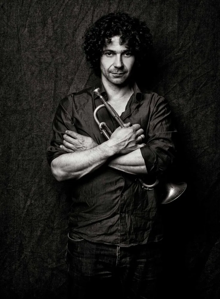 Black and white photo of a curly-haired man with his arms crossed in front of his chest, holding a trumpet in one hand.