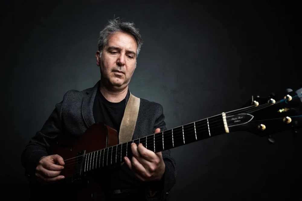 A man with short gray hair in a gray sacko plays the guitar and looks at the guitar neck.