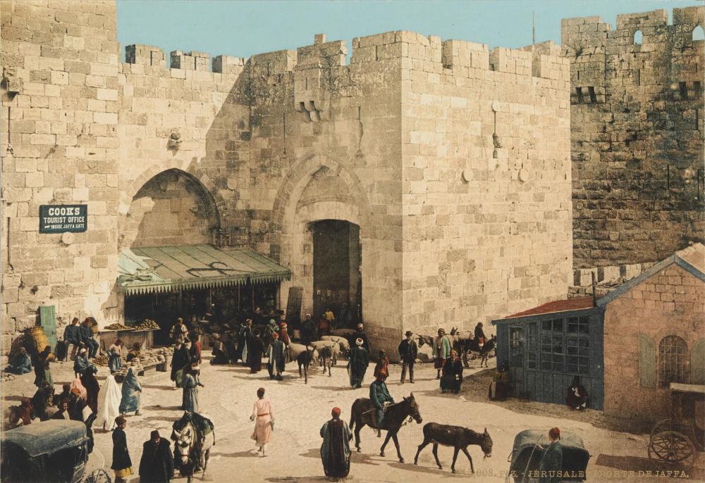 Die kolorierte Fotografie zeigt die massive Stadtmauer von Jerusalem. Auf dem Vorplatz des Jaffa-Tors sind viele Menschen unterwegs, zudem sind Marktstände, Esel und ein Auto zu sehen