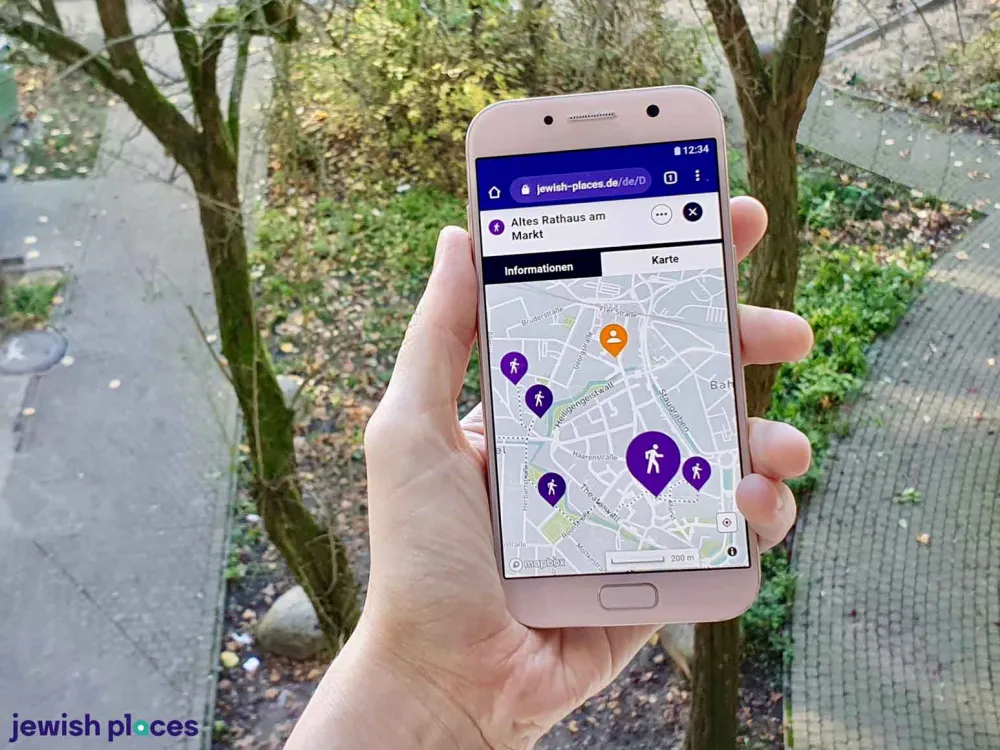 One hand holds a cell phone up to the camera. A map application can be seen on the screen. A bed with shrubs and paths can be seen in the background.