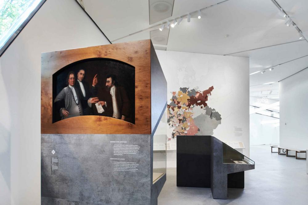 Showcases made of concrete and wood, in them a painting with three persons in the background a map of the territory of present-day Germany