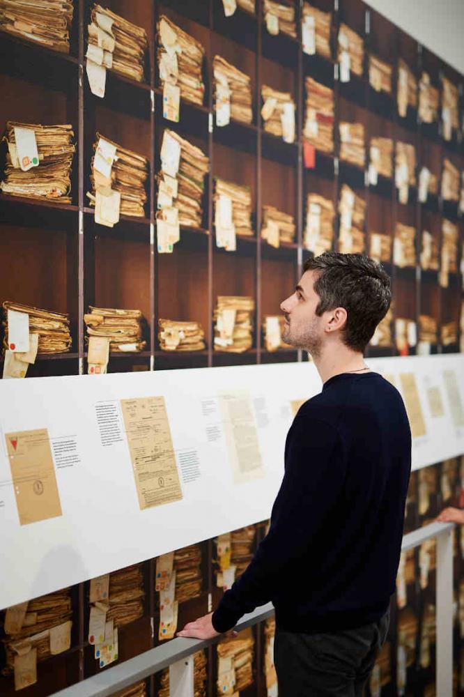 Man stands in front of a wall with a photo of a shelf with many old files