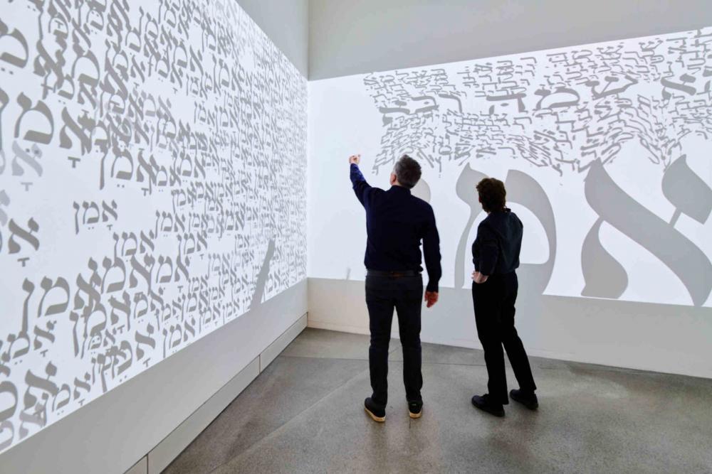 Große Leinwand mit hebräischen Buchstaben vor der zwei Personen stehen.