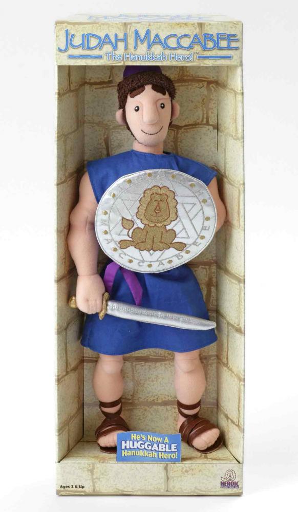 A Judah the Maccabee doll in its original sales packaging. The figure is smiling, dressed in a sleeveless knee-length blue tunic and Roman sandals, and holds a sword and a shield emblazoned with a lion and a Star of David