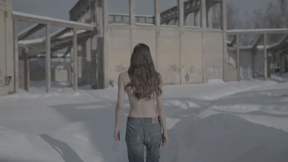 Screenshot of a long-haired person with jeans and bare upper body from behind, walking through snow towards a building.