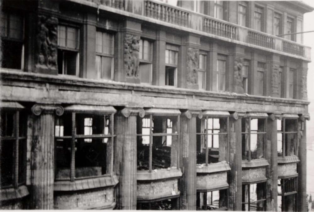 Das Foto zeigt ein Gebäude mit zerstörten Fensterscheiben und die Verwüstung im Inneren der ersten beiden Stockwerke des Kaufhauses. Das Foto ist auf Höhe des 2. Stockwerks, vermutlich vom gegenüberliegenden Gebäude aus, aufgenommen worden.