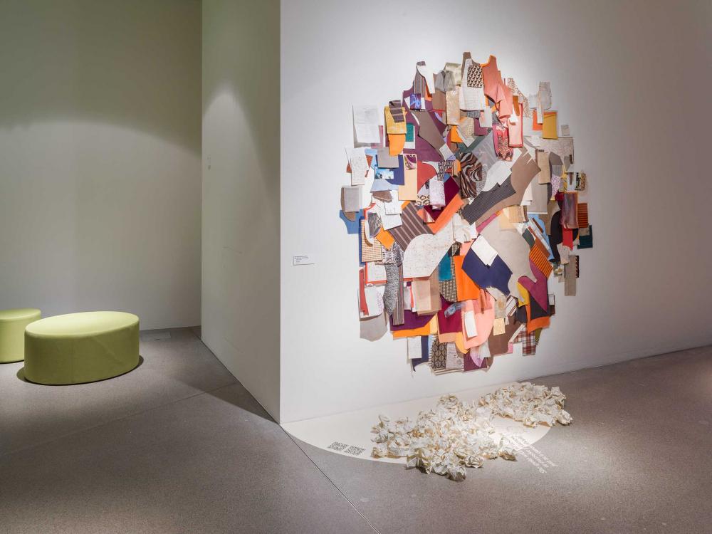 View of the exhibition space: On the wall hangs a work of art consisting of colorful scraps of fabric and written notes. In front of it, crumpled pieces of paper lie on the floor; on the left of the picture are two green poufs.