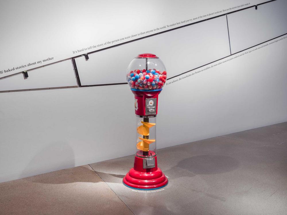 Exhibition view showing a large, free-standing gumball machine. Its round, glass container is filled about halfway with colorful balls.