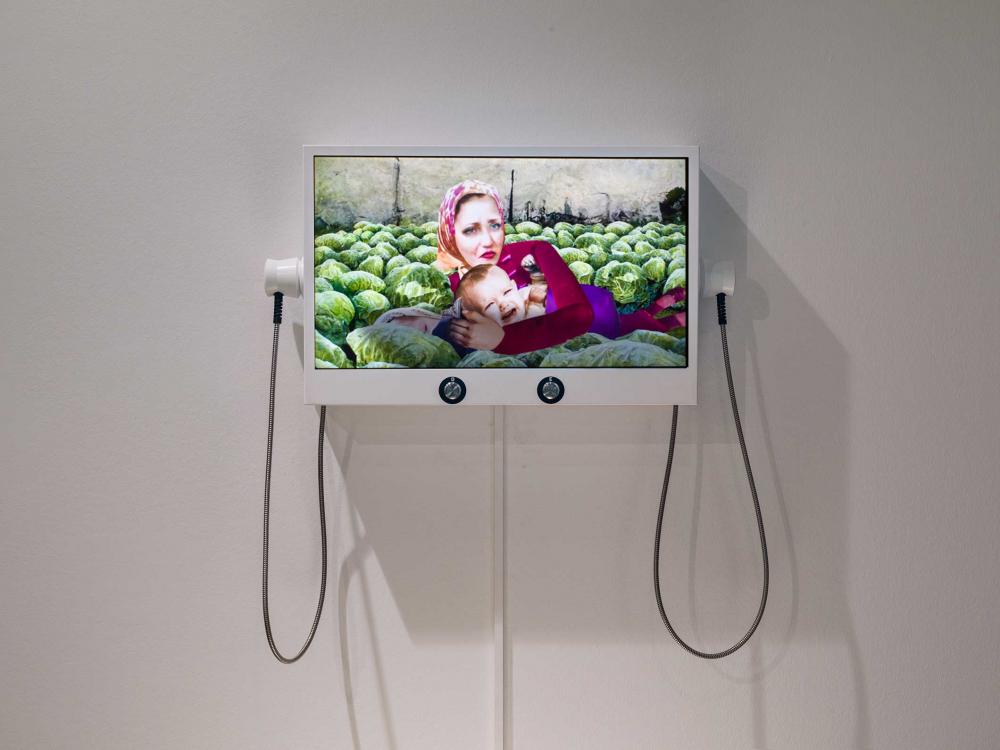 A monitor hangs on the exhibition wall. It shows a film still of a woman with a crying baby and another child. All three are lying between cabbage heads in a field.