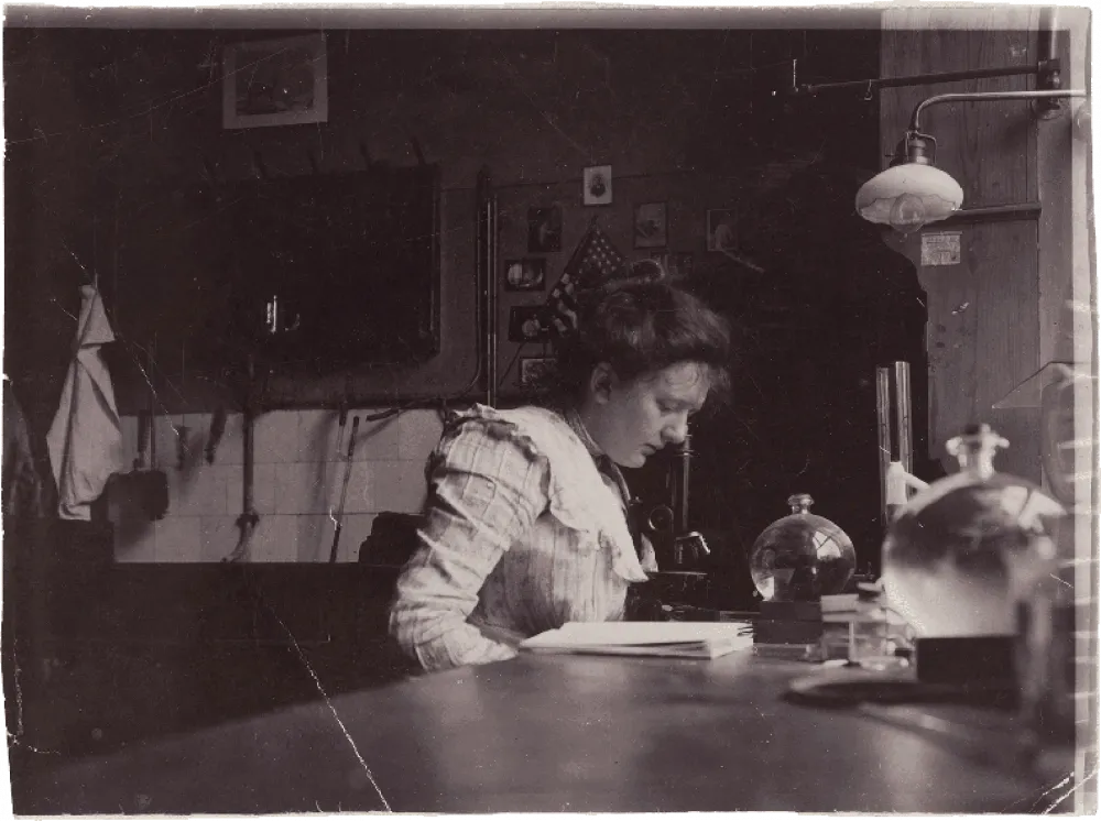 Historical black and white photograph of a young woman reading bent over a table.