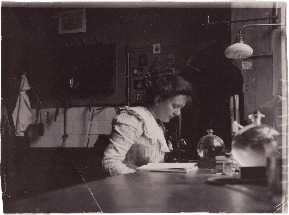 Historical black and white photograph of a young woman reading bent over a table.