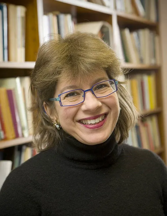 A blonde woman with blue glasses and a black turtleneck sweater stands in front of a bookshelf and smiles into the camera.