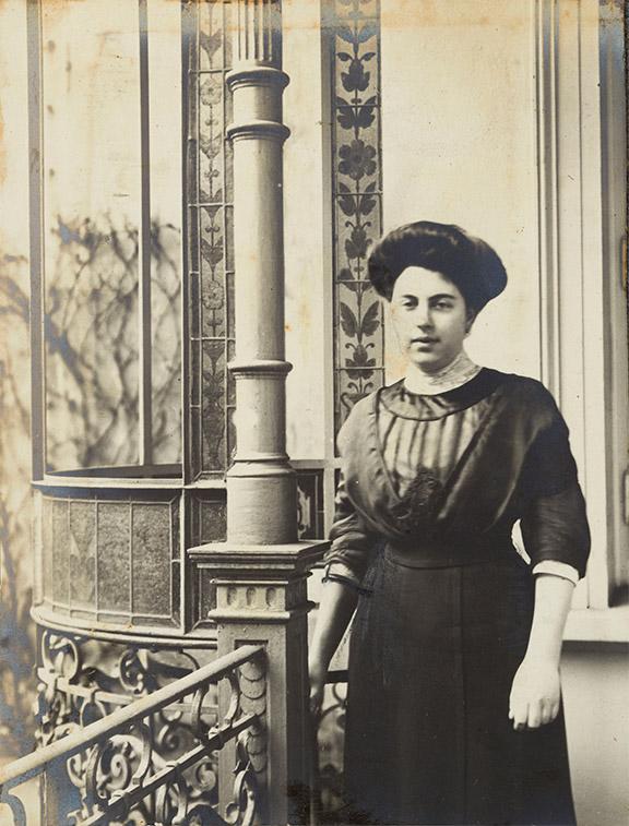 Black and white portrait photo of a young woman. She is wearing a high-necked dress in the style of the 1910s and an updo and is standing in front of a house whose magnificent facade decoration can be seen in the background.