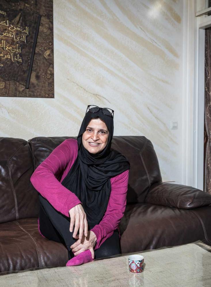 A woman with a black headscarf and a pink top is sitting on a leather couch and smiling.