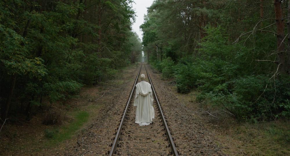The film still shows a person from behind, dressed in a white frock. The person follows railroad tracks that run dead straight through a forest