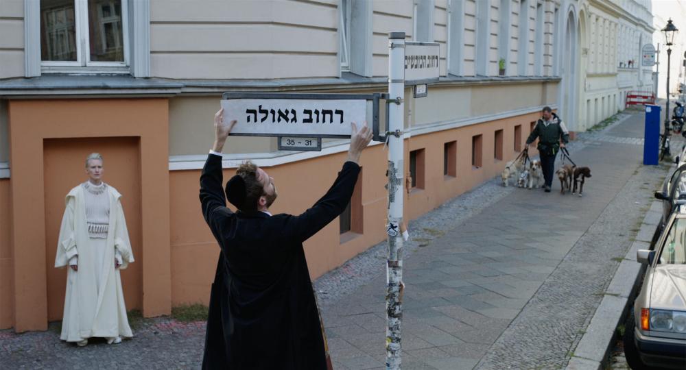 Ein Mann mit Kippa montiert ein Straßenschild mit hebräischem Straßennamen. Links von ihm steht eine Frau in weißen Gewändern im Rahmen einer zugemauerten Tür, die Teil des orangefarbigen Sockels eines renovierten Altbaus ist, und schaut ihm zu
