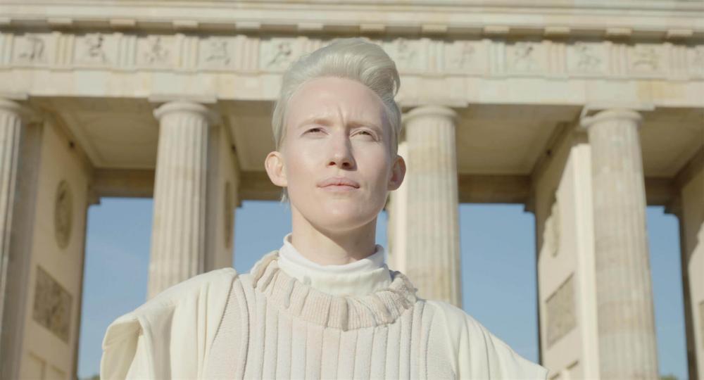 Bust portrait of an androgynous looking platinum blonde person with short hair and white top. Behind her are the columns of the Berlin Brandenburg Gate against a blue sky