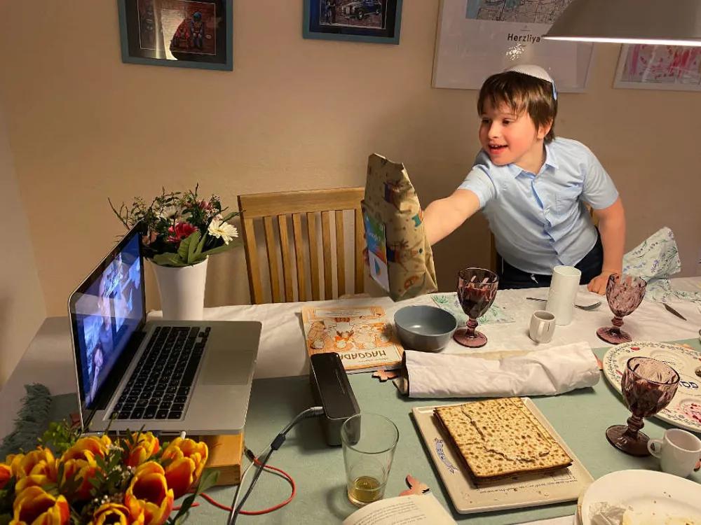 A boy with kippah holds a brown paper bag into the camera from a laptop.