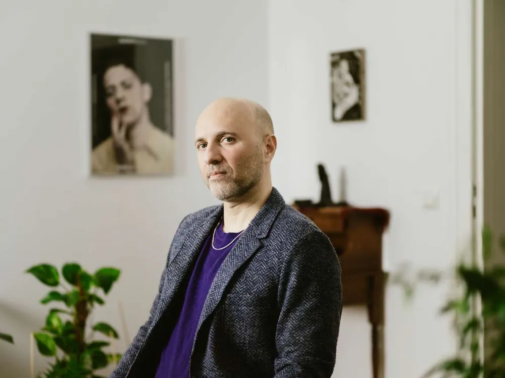 Ein Mann mit Dreitagebart, Glatze und Fischgrätenjackett über lila Shirt und Kette sitzt in einem Raum mit Pflanzen und Bildern an den Wänden und schaut mit lebhaftem Blick in die Kamera.