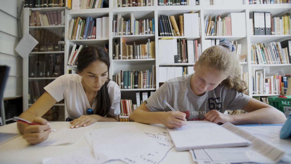 Two young people sit at a table and write with pens in a notebook behind a shelf at the library of the Steglitz Museum.