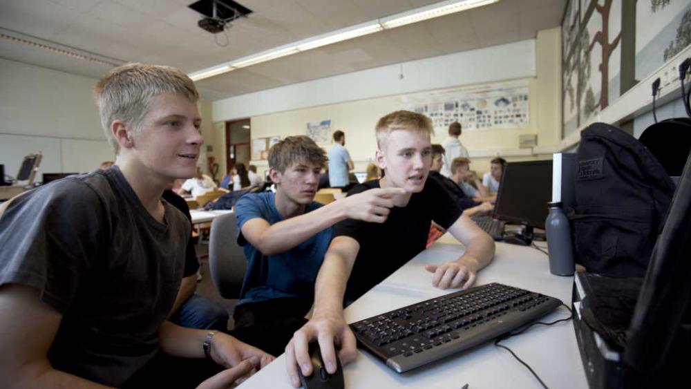 Young people sit in a large room. Three young people sit in front of a keyboard in the foreground.