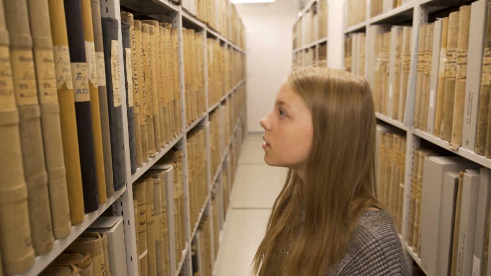 A girl stands in a room in the Brandenburg Main State Archive between shelves of old books. 